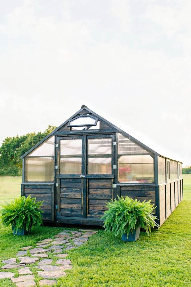 Exterior view of a gray stain Yoderbilt Greenhouse with double doors that are closed. Two potted plants sit outside the doors.