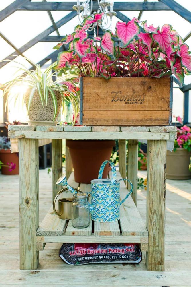 Close up of a wooden table with a wooden box with flowers in it, water can and potted plants. It's sitting inside a Yoderbilt Greenhouse.