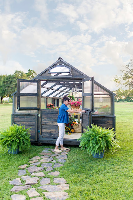 Angela Yoder standing in front of a Yoderbilt Greenhouse with gray stain. The top windows are open and the bottom right door is also open. Inside you can see various plants.