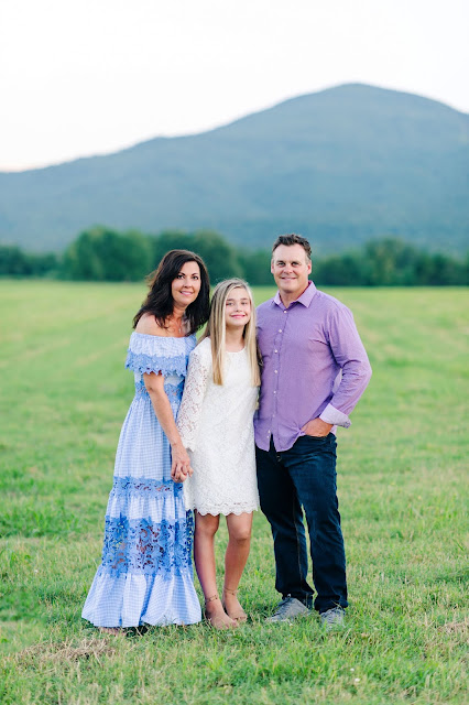 Angela, Travis, and daughter standing in a field with a mountain in the background posing for a picture.