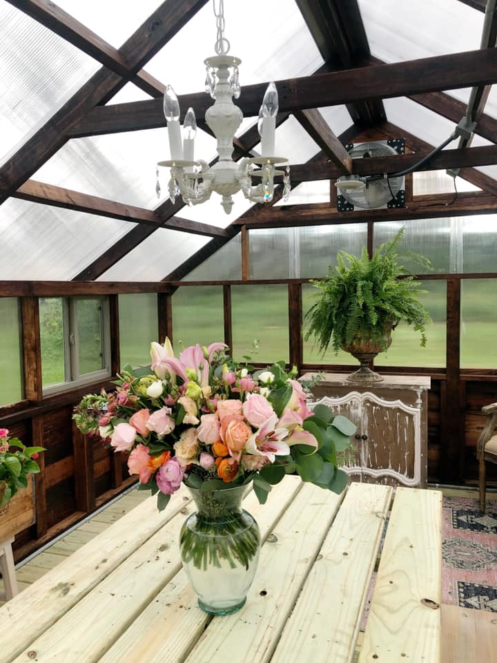 Interior view of a Yoderbilt Greenhouse with brown stain. Inside there is a wooden table with a vase of flowers on it and a chandelier hanging from the roof. Also a chair in the corner.