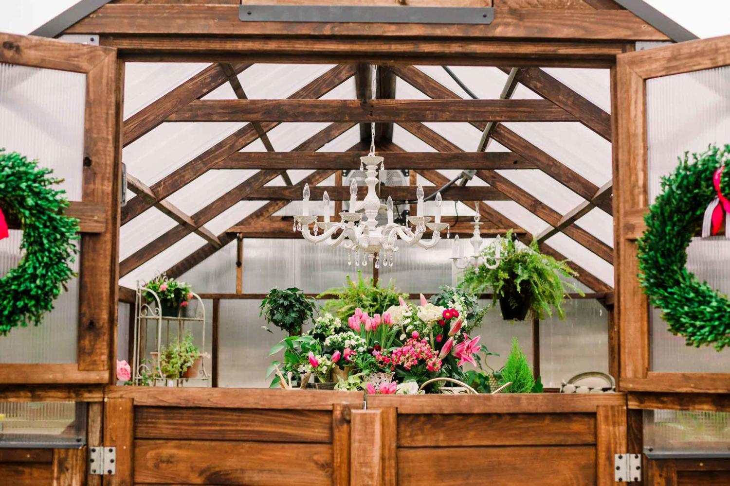 Close up of the interior of a Yoderbilt Greenhouse with brown stain. The bottom double doors are close but the top are open. Inside is various plants and flowers with a chandelier hanging from the roof.
