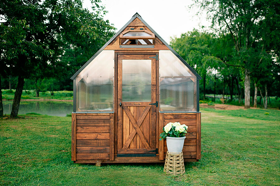 Front view of a 8x20 Yoderbilt Legacy Greenhouse with brown stain. The single door closed. One potted plant is outside by the door.