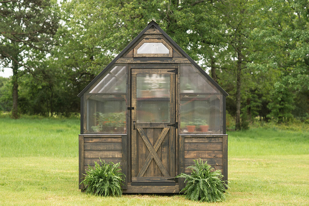 Front view of a 8x12 Yoderbilt Legacy Greenhouse with gray stain. The single door closed with plants on both sides of the door.