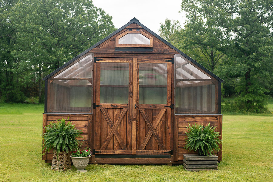 Front exterior view of a 12x16 Yoderbilt Legacy Greenhouse with brown stain. Planters on both sides of the closed double doors.