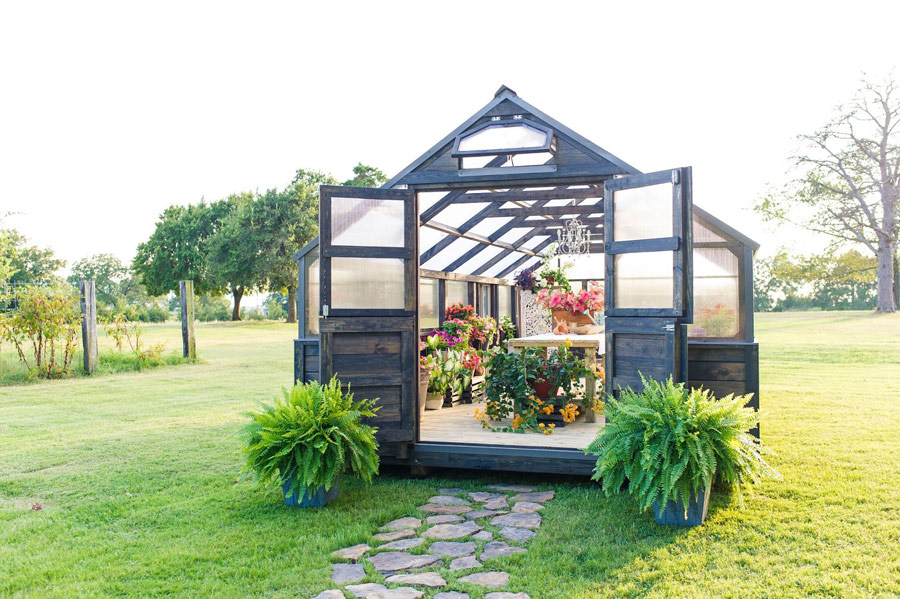Exterior view of a gray stain Yoderbilt Greenhouse with double doors that are held open by two potted plants. The interior can be seen with a wooden table and several plants.