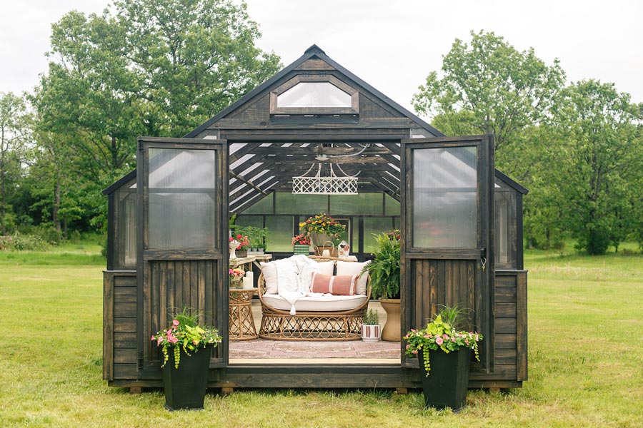 Front view of a Yoderbilt Greenhouse with gray stain. Double doors are held open by potted plants. There are various plants spread around on wooden tables. In front is a white couch with a side table and large potted plant next to it.