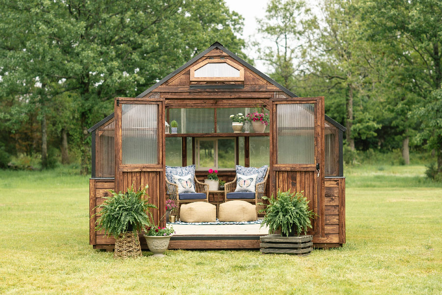 Front view of a Yoderbilt Greenhouse with brown stain. Double doors are held open by planters. There are two blue chairs and side table sitting inside. Plants are on the back shelf of the Greenhouse.