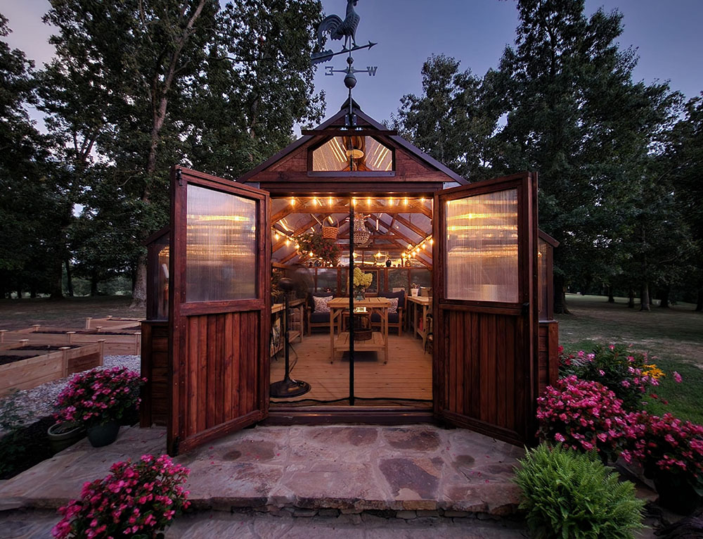 Exterior of a Yoderbilt greenhouse at night with brown stain and brown metal trim. The double doors are open and lights are strung inside.