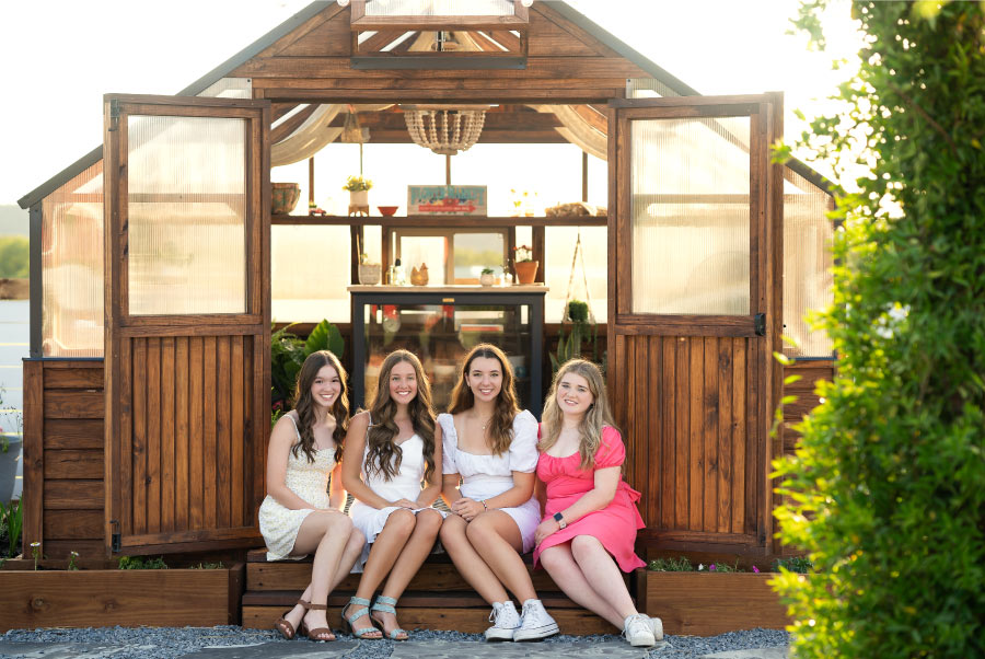 Four girls smiling in front of a Yoderbilt greenhouse with brown stain.