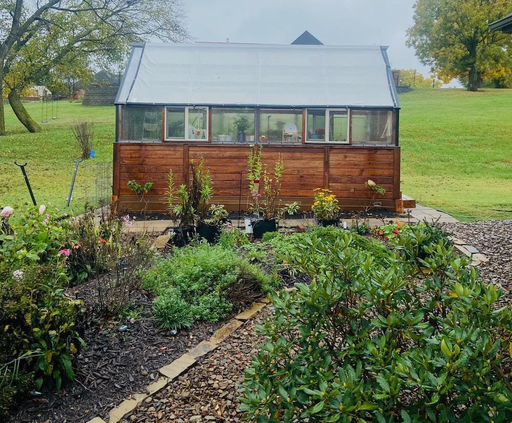 Side view of a Yoderbilt Greenhouse with brown stain with two windows. In front is a garden with various plants.
