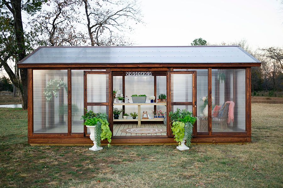 Side view of a Yoderbilt Greenhouse with brown stain. The side doors are held open by two potted plants. Inside there is a wooden bench with various plants and object on it.