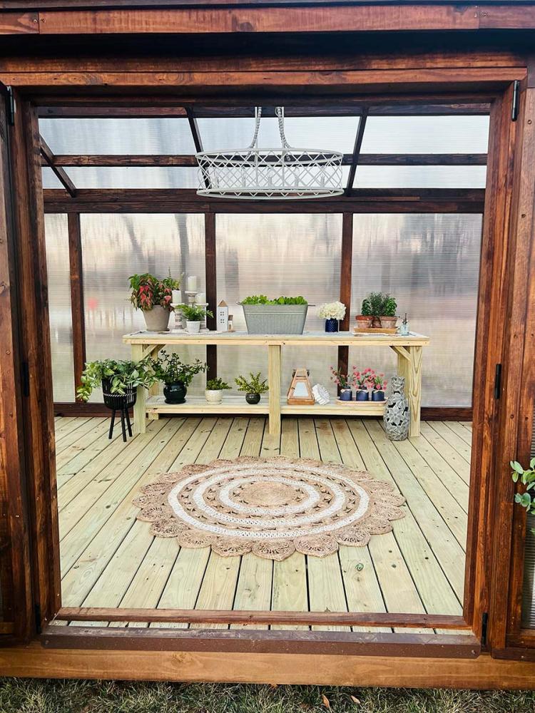 12x20 Traditional Yoderbilt Greenhouse with brown stain exterior close-up of the open double door. Inside, a rug and gardening table filled with plants.