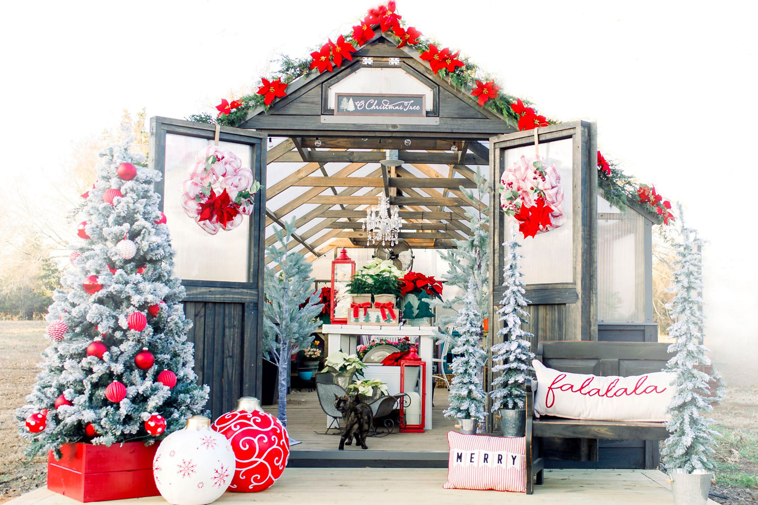 Exterior of a Yoderbilt Greenhouse with holiday decor. Poinsettia garland lines the roof and the deck on front features a Christmas tree, large ornaments, and a bench.