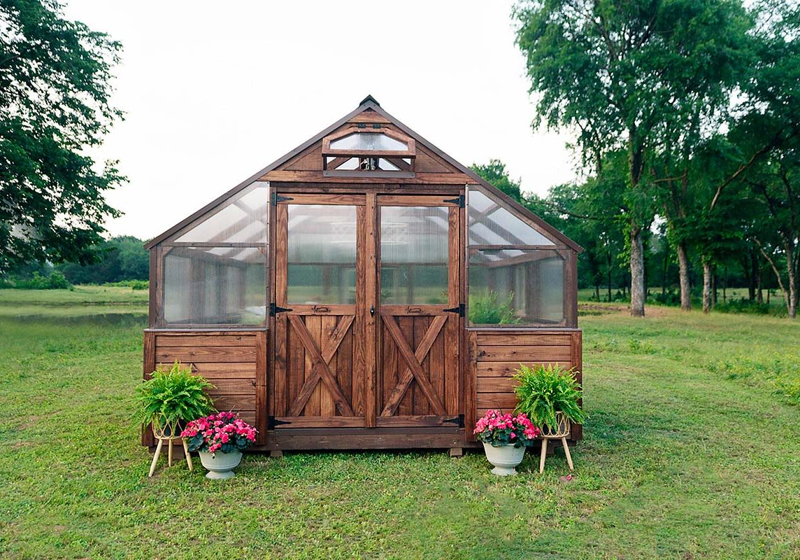 Exterior front view of a 12x12 Yoderbilt Legacy Greenhouse with brown stain. Planters on both sides of closed double doors.